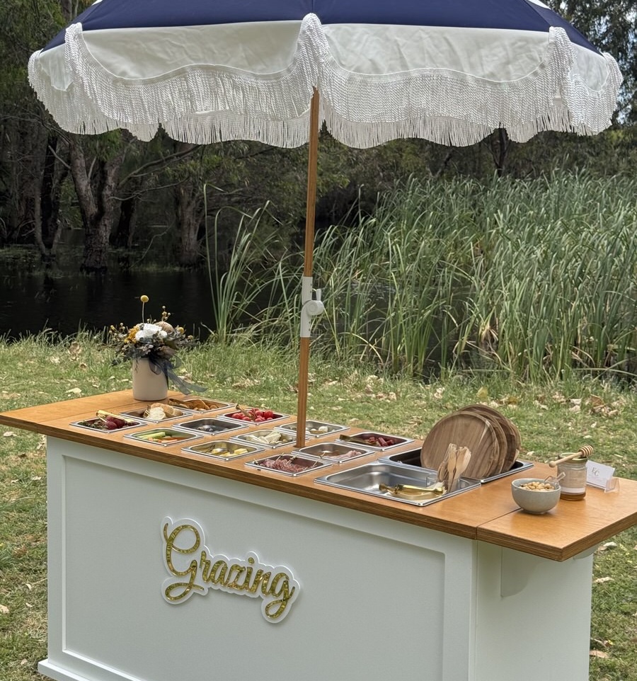 Charcuterie grazing cart fully loaded with food, timber top, fringed parasol and gold lettering